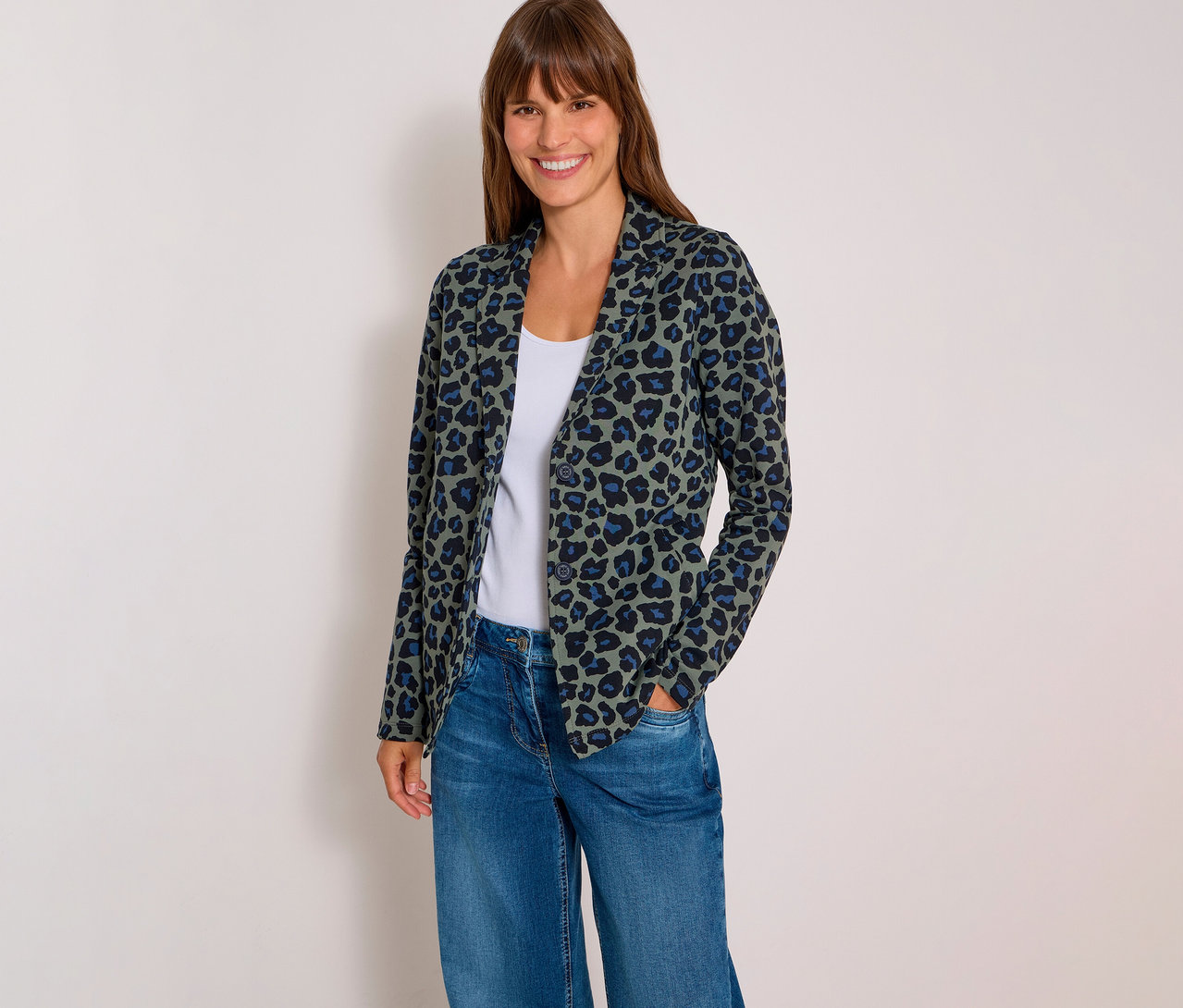 Femme souriante portant un blazer en jersey vert à motif léopard, un débardeur blanc et un jean bleu, les mains dans les poches.