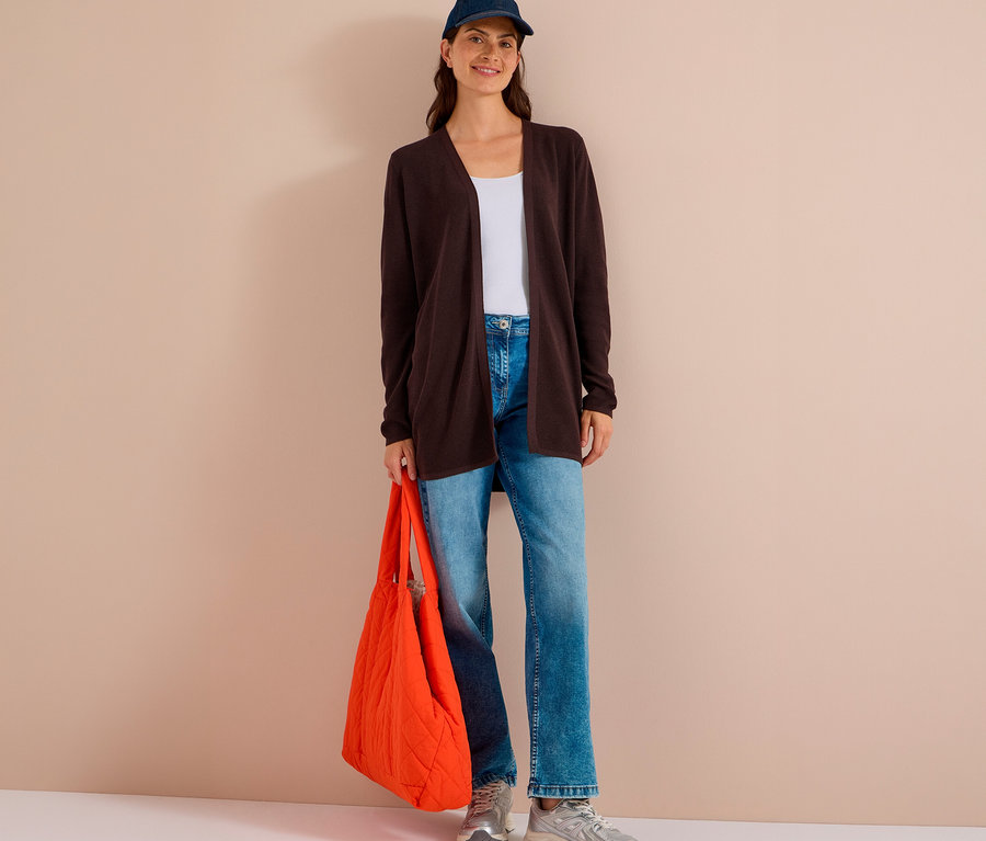 Femme souriante debout, portant un cardigan CECIL marron foncé, un top blanc, un jean, une casquette bleue et tenant un sac matelassé orange.