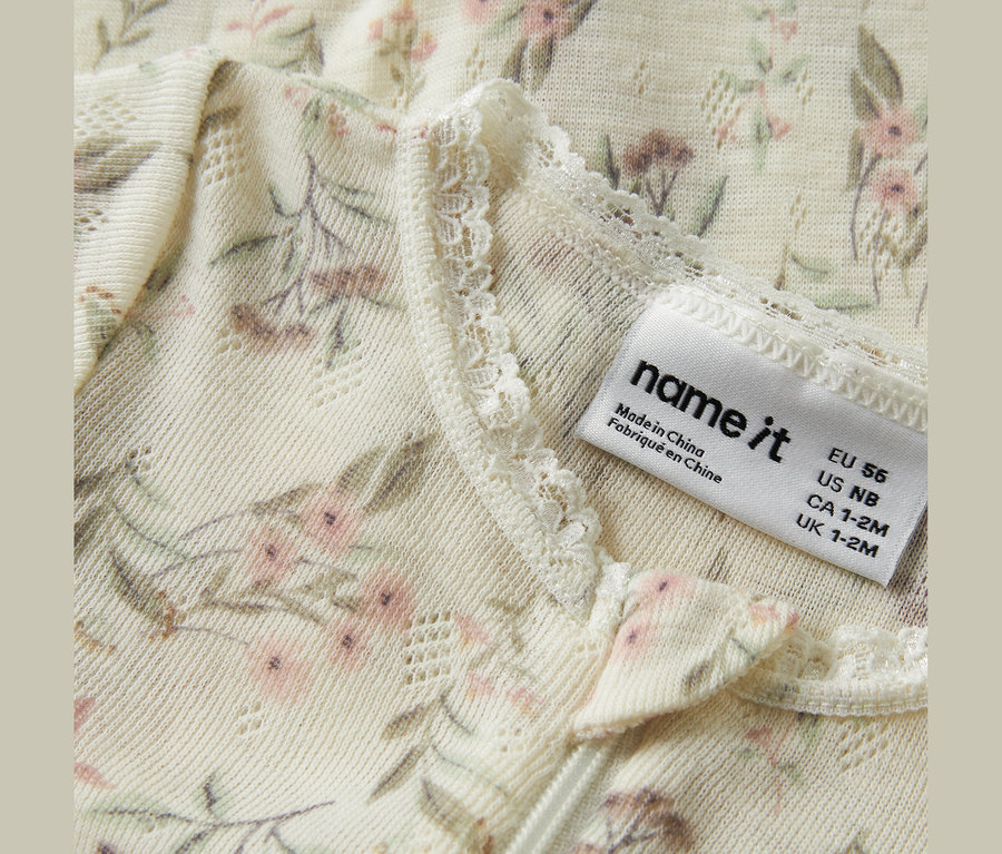 Vêtement couleur crème avec un délicat motif floral de fleurs roses et de feuilles vertes, présentant une texture tricotée avec de petits trous et une bordure en dentelle. Une étiquette de marque 'name it' indique 'Made in China' et les tailles EU 56, US NB, CA 1-2M, UK 1-2M.