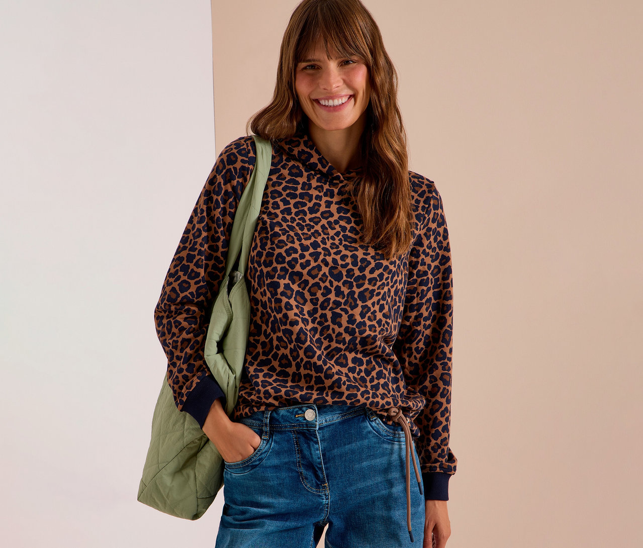 Femme souriante portant un sweatshirt CECIL à imprimé léopard marron et noir et un jean, avec un sac matelassé vert sur l'épaule.