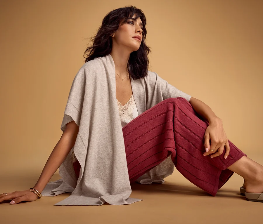 Femme assise sur un sol marron clair, portant un poncho gris clair sur un débardeur en dentelle blanche, un pantalon large rouge à rayures horizontales et des chaussures plates gris clair. Elle regarde vers la droite, main gauche au sol, main droite sur sa jambe.