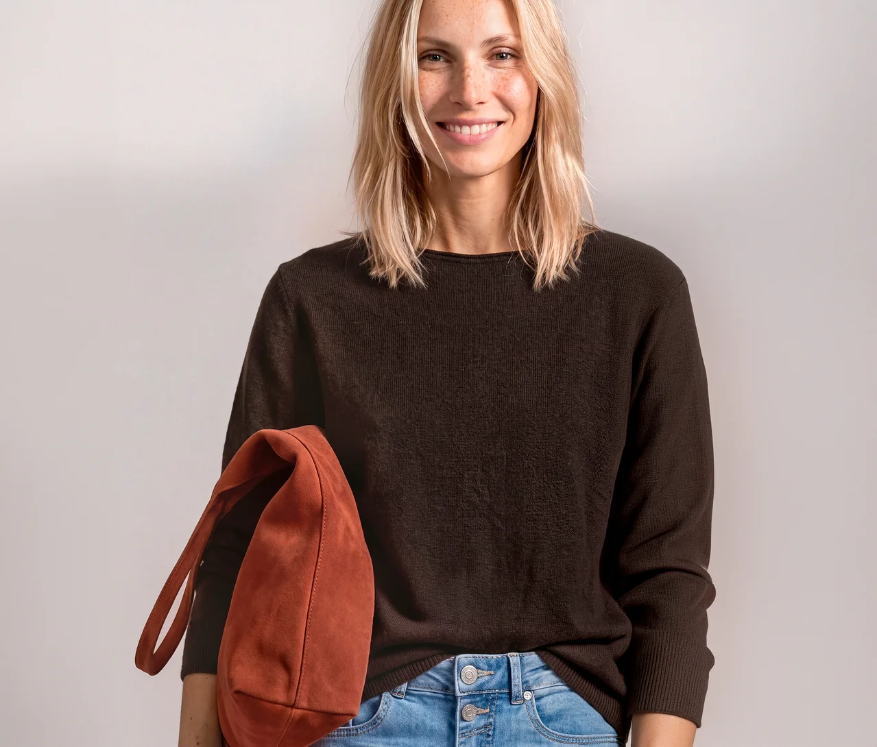 Femme souriante portant un pull en maille Street One marron et un jean bleu clair, tenant un sac marron-rouge.
