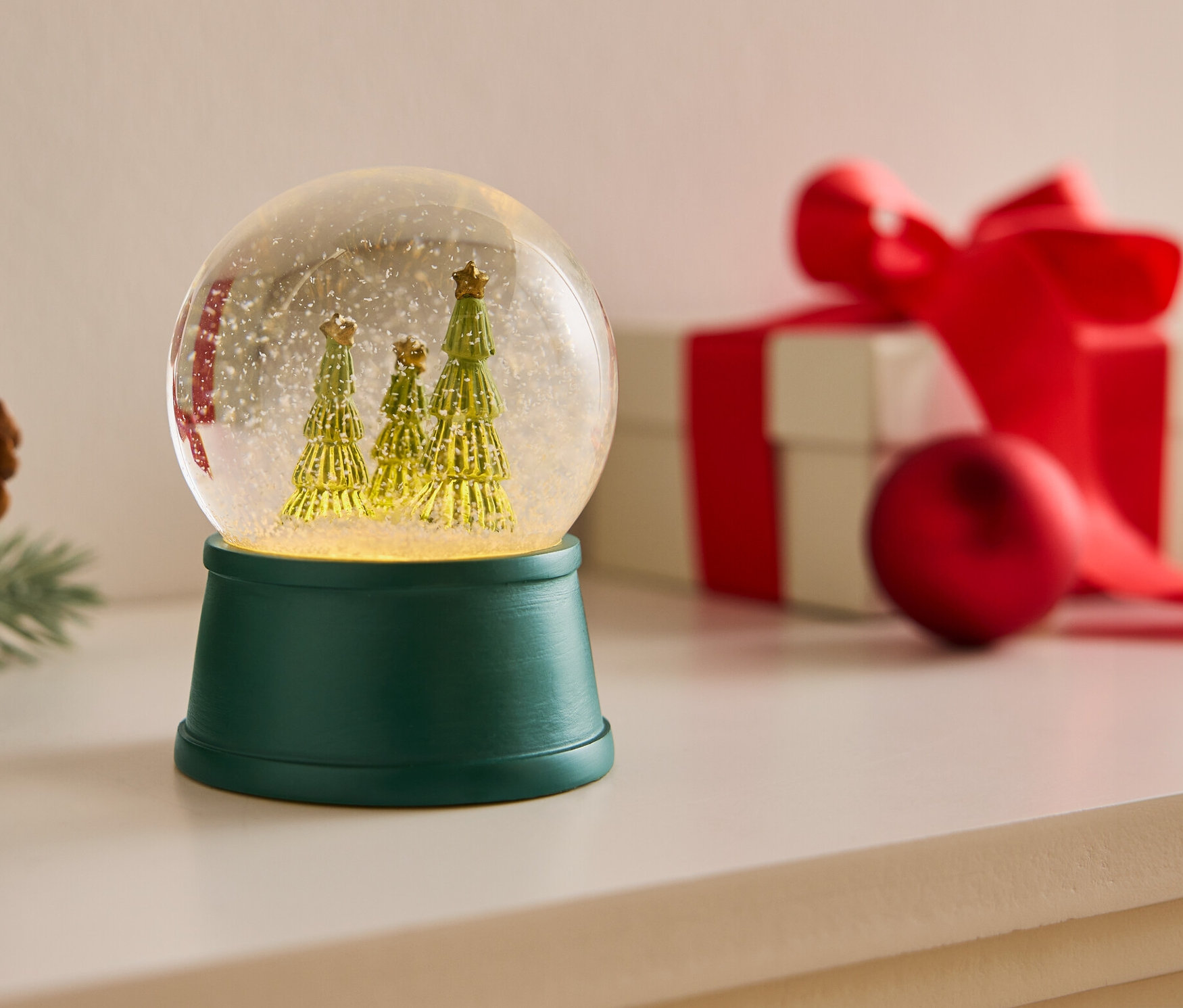 Boule à neige avec trois arbres sur une base verte à côté d'une pomme de pin et d'un cadeau avec un ruban rouge.