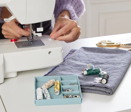 Femme enfilant du fil dans une machine à coudre. Kit de fil à coudre dans une boîte bleue sur la table.