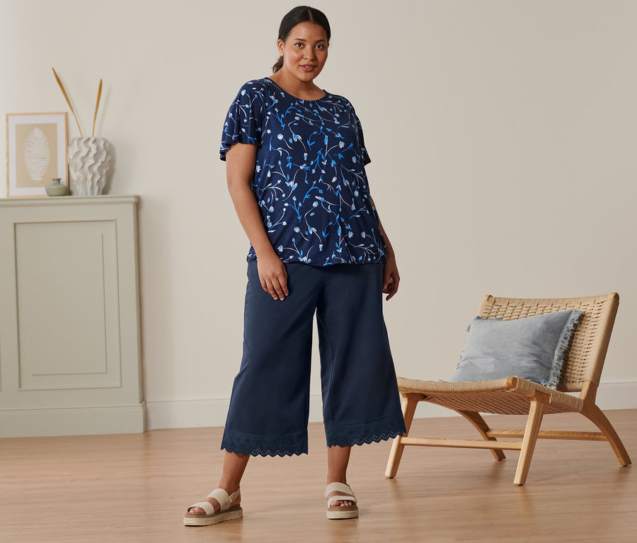 Une femme debout porte un chemisier bleu foncé à manches papillon, une jupe-culotte à broderie et des sandales à plateau en cuir.
