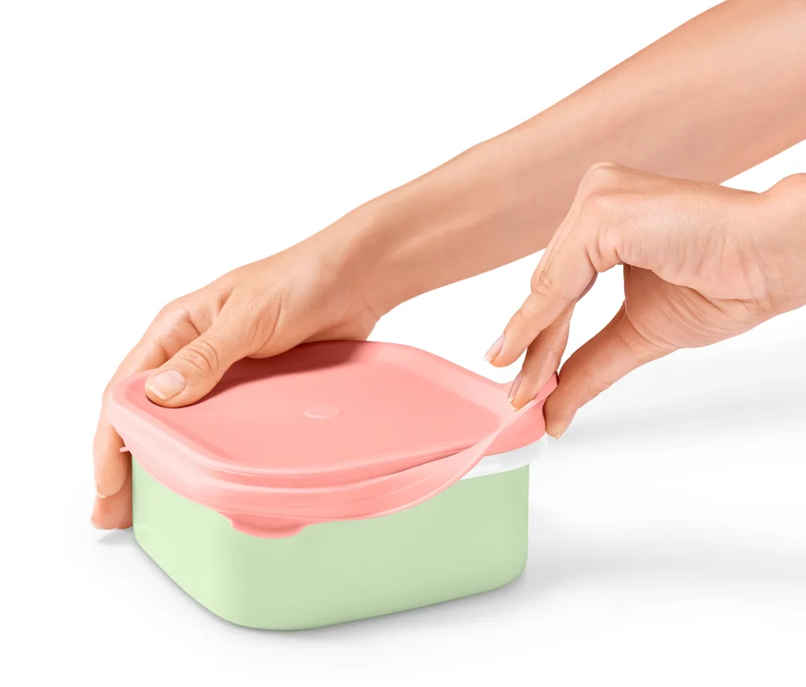 Des mains ouvrent une boîte à lunch émaillée avec un couvercle rose et un fond vert.