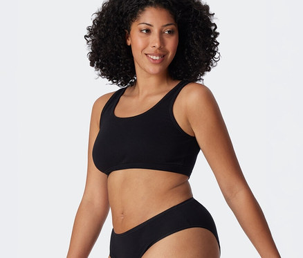 Femme souriante aux cheveux bouclés foncés portant une brassière SCHIESSER noire et une culotte noire.