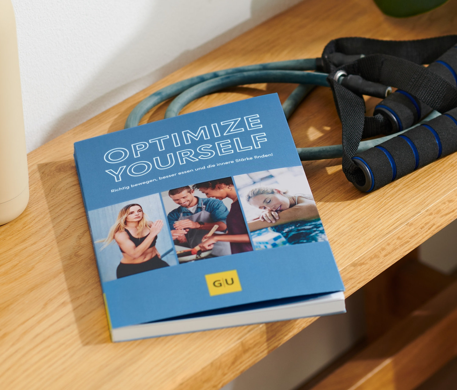 Sur un banc en bois se trouvent le livre « Optimize Yourself », une bouteille d'eau beige et un expandeur.