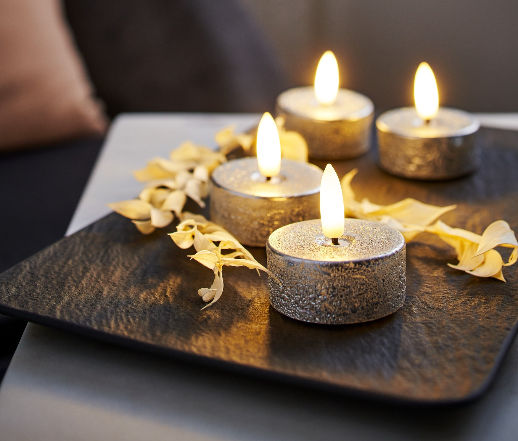 Quatre lampes à thé LED en cire argentées présentées sur un plateau noir et décorées de fleurs séchées.