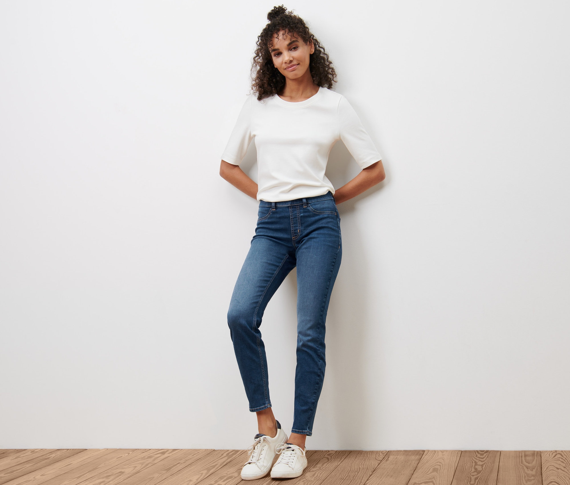 Une femme pose sur un plancher en bois en tregging bleu « Fit Nelly » et un t-shirt blanc.