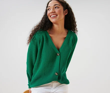Femme aux cheveux bouclés portant un cardigan vert en grosse maille et un pantalon blanc.