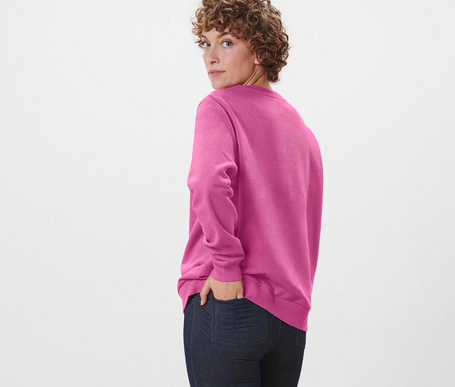 Une femme aux cheveux bouclés regarde par-dessus son épaule et pose en portant un sweatshirt rose vif.