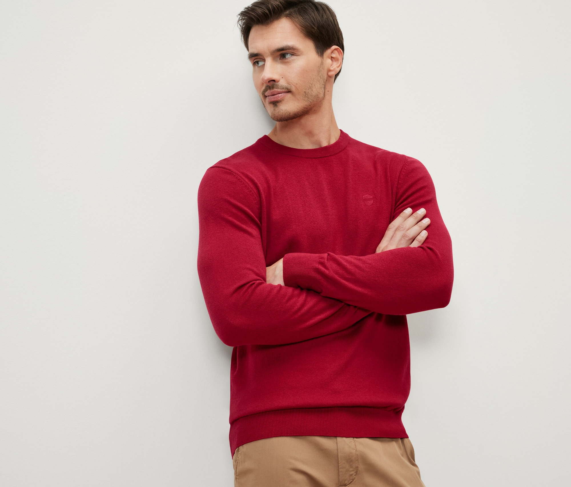 Un homme, les bras croisés, porte un pull rouge à encolure ronde.
