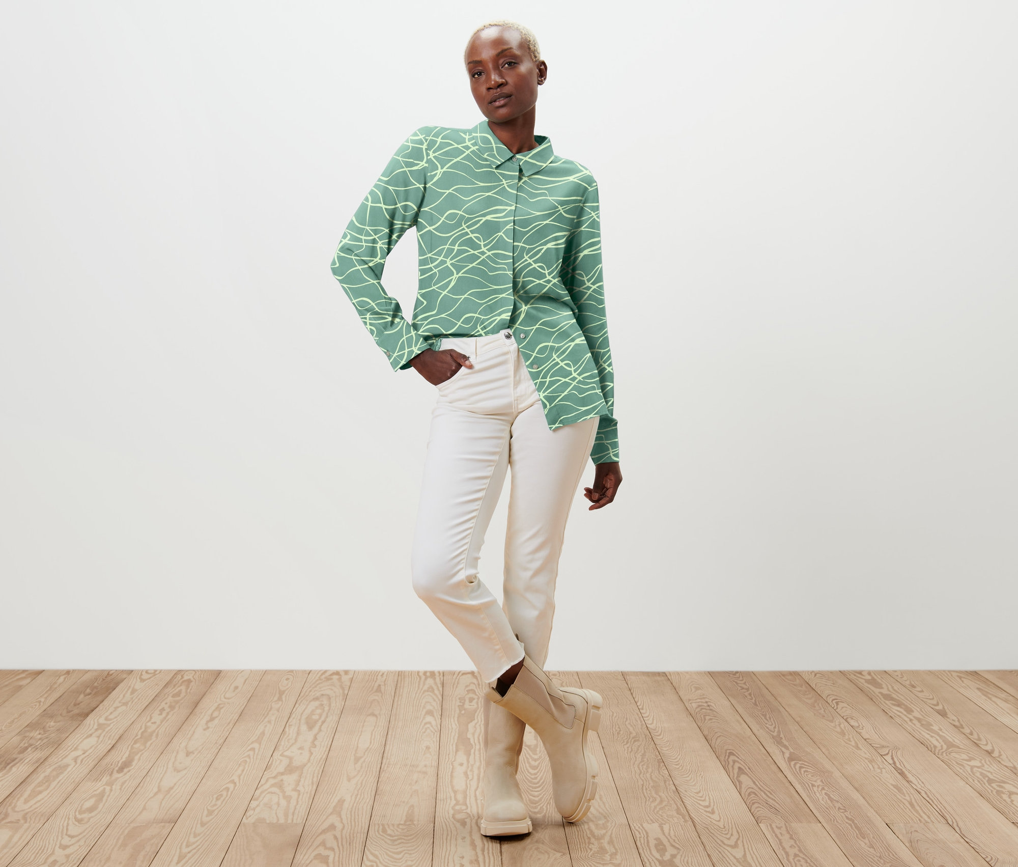 Femme posant en chemisier imprimé vert, jean blanc et bottes beiges.