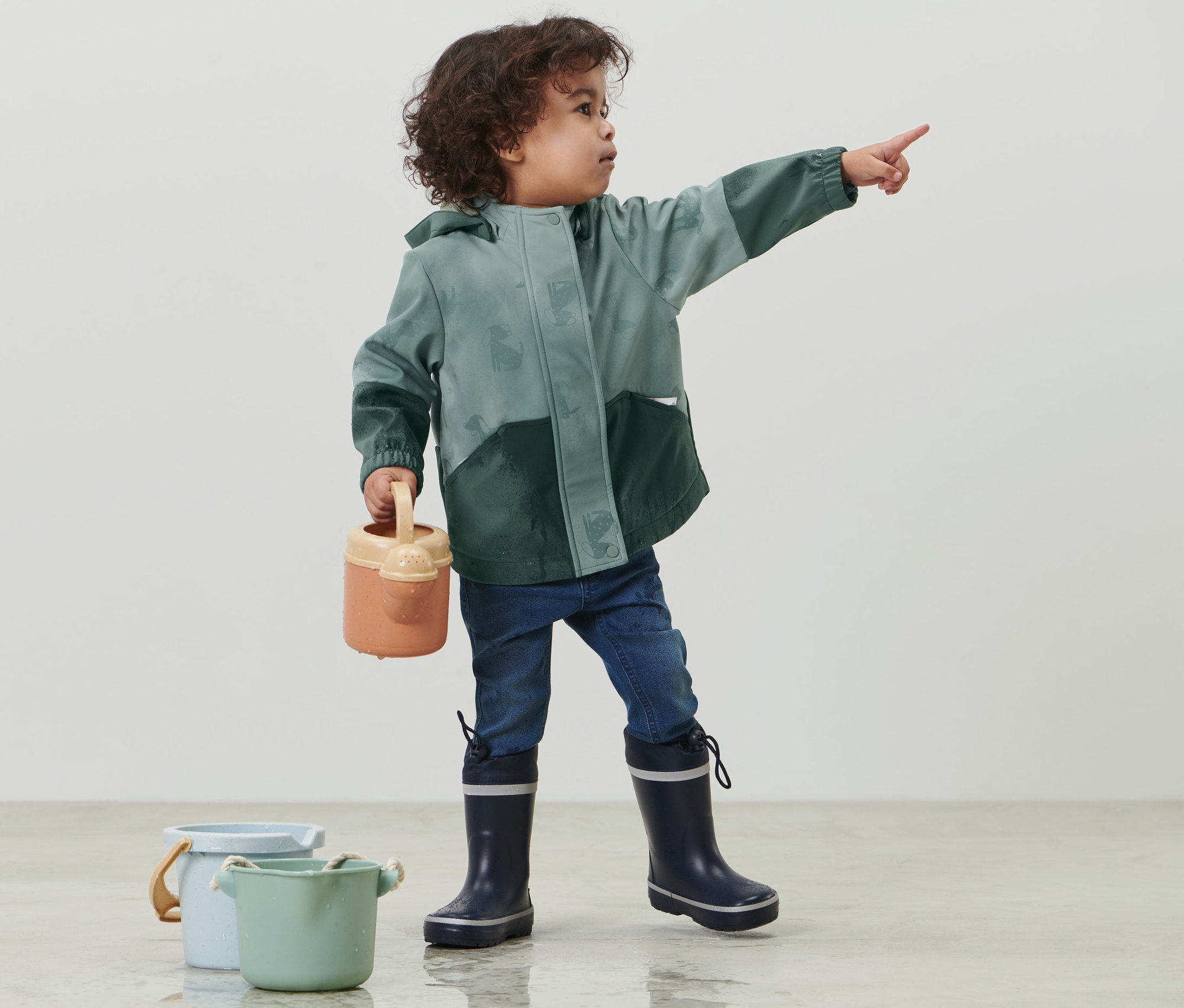 Garçon portant une veste en softshell pour enfant qui change de couleur quand il pleut, vert, pointant du doigt, tenant un arrosoir, avec deux seaux à côté de lui.