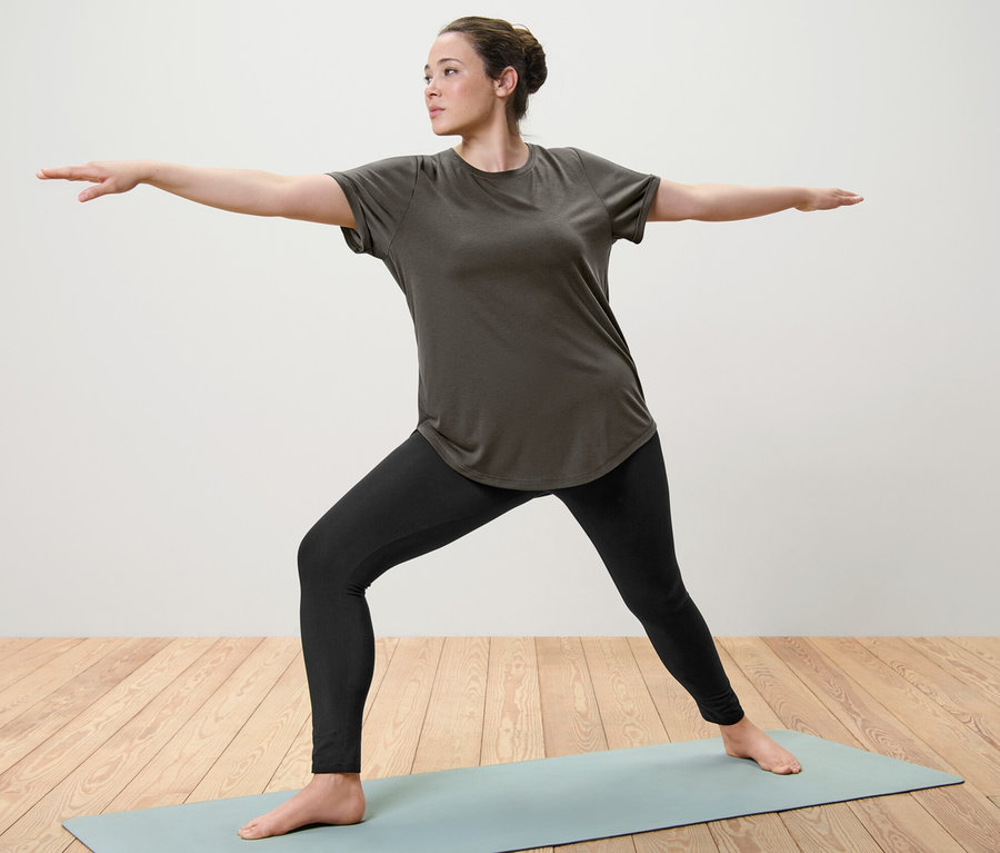 Femme faisant du yoga sur un tapis, portant un t-shirt long vert olive et un collant de sport.