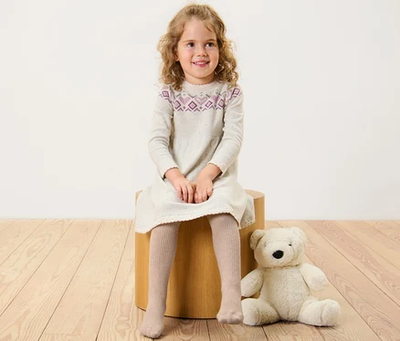 Une fille est assise sur un tabouret en bois, portant une robe et des collants pour enfant avec laine RWS.