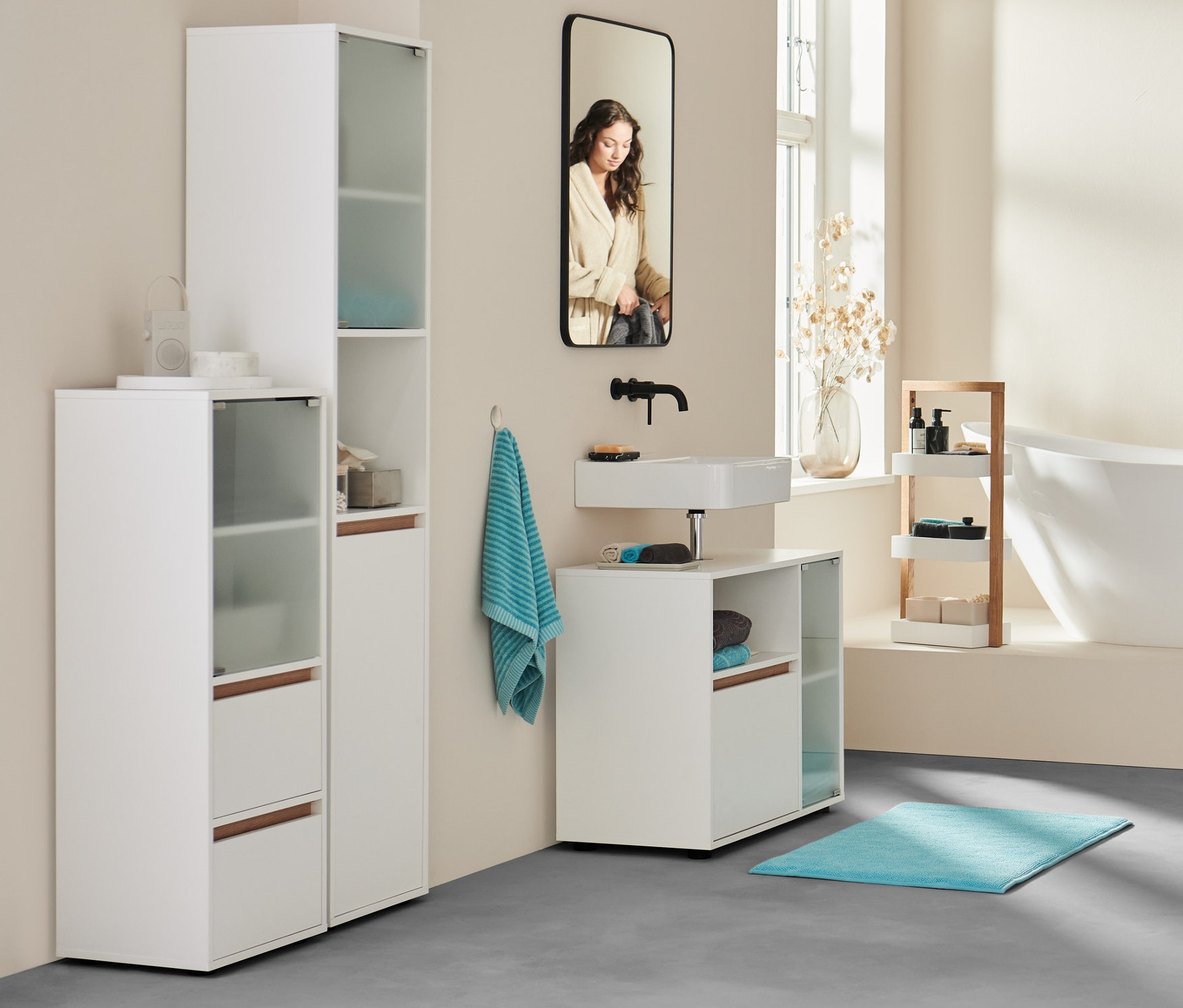 Dans une salle de bain, une femme en peignoir se tient près de la baignoire. Au premier plan, buffet de salle de bain, drap de bain extra grand anthracite et 2 serviettes turquoise.