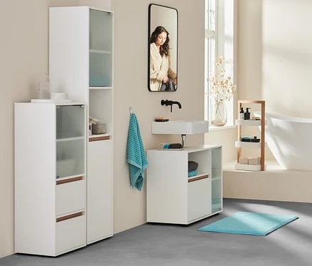 Dans une salle de bain, une femme en peignoir se tient près de la baignoire. Au premier plan, buffet de salle de bain, drap de bain extra grand anthracite et 2 serviettes turquoise.