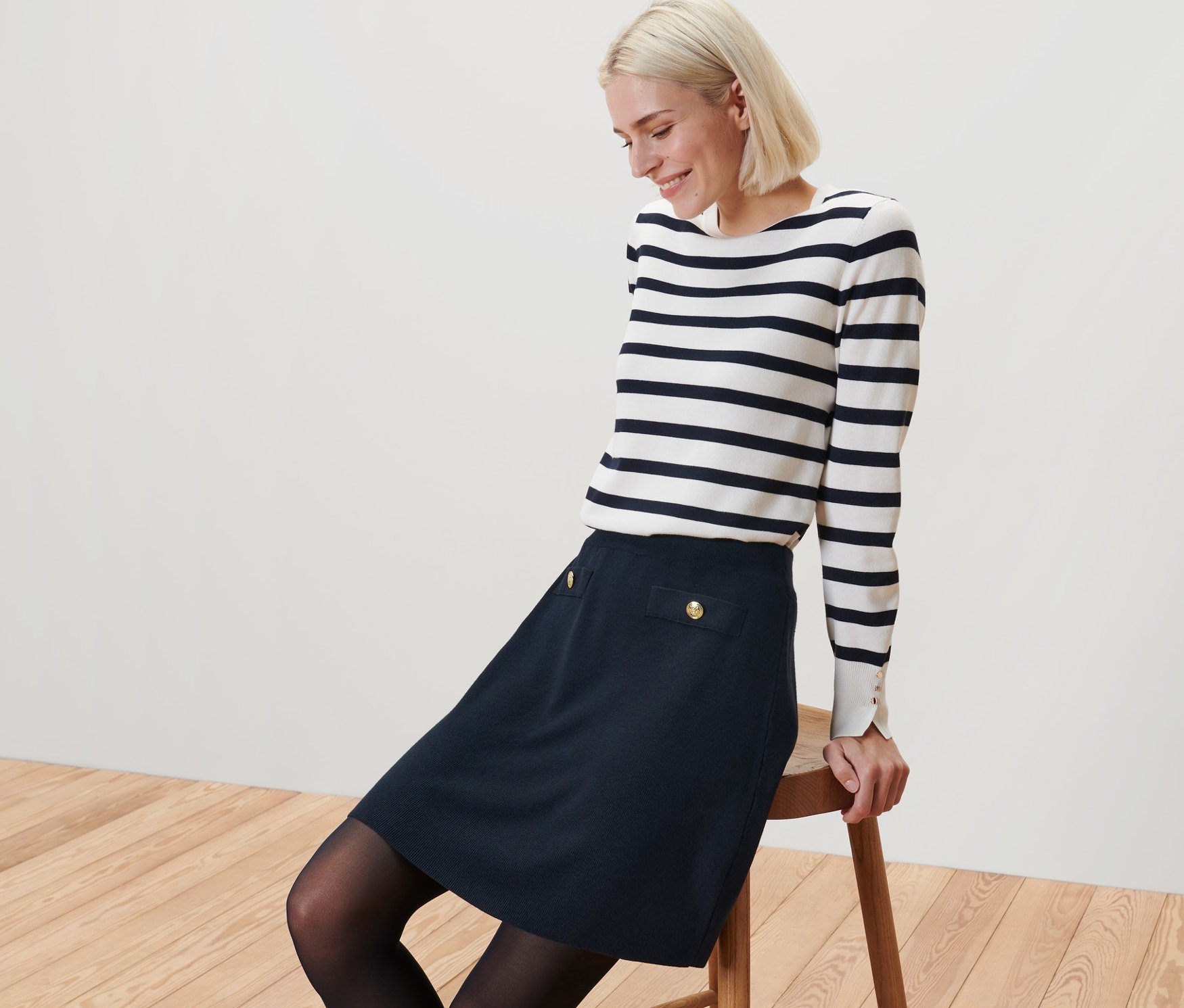 Femme blonde assise sur un tabouret portant une jupe en maille avec boutons décoratifs et un haut rayé.