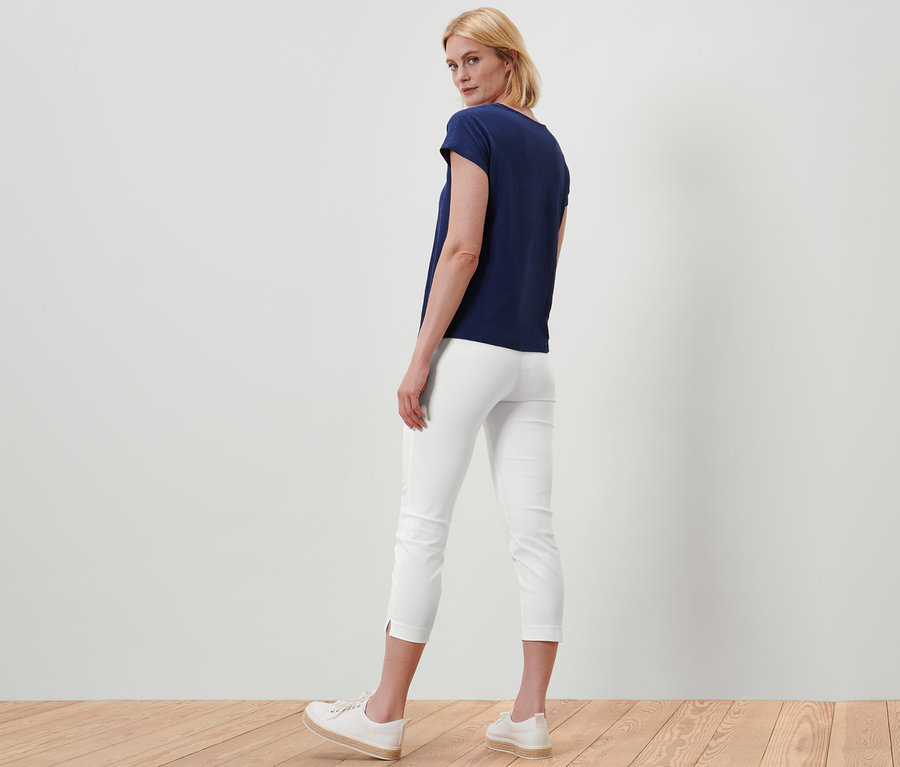 Une femme se tient dos à la caméra et porte un pantalon stretch 7/8 blanc et un t-shirt bleu marine.