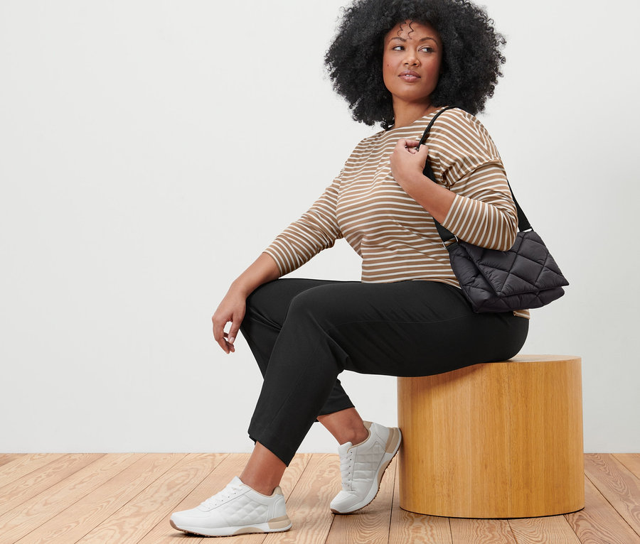 Une femme est assise sur un tabouret, portant un pantalon de jogging noir, un t-shirt manches longues à rayures et un sac matelassé à bandoulière.