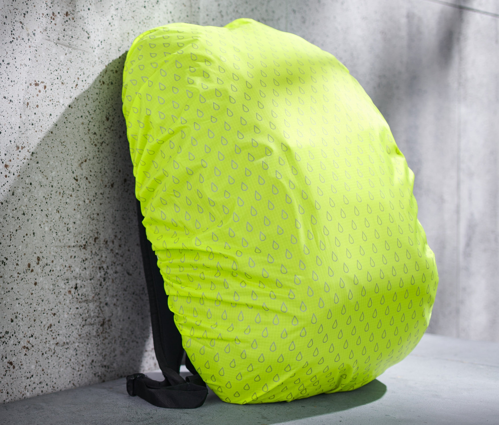 Housse de protection jaune pour sac à dos avec motif de gouttes sur un sac à dos appuyé contre un mur.