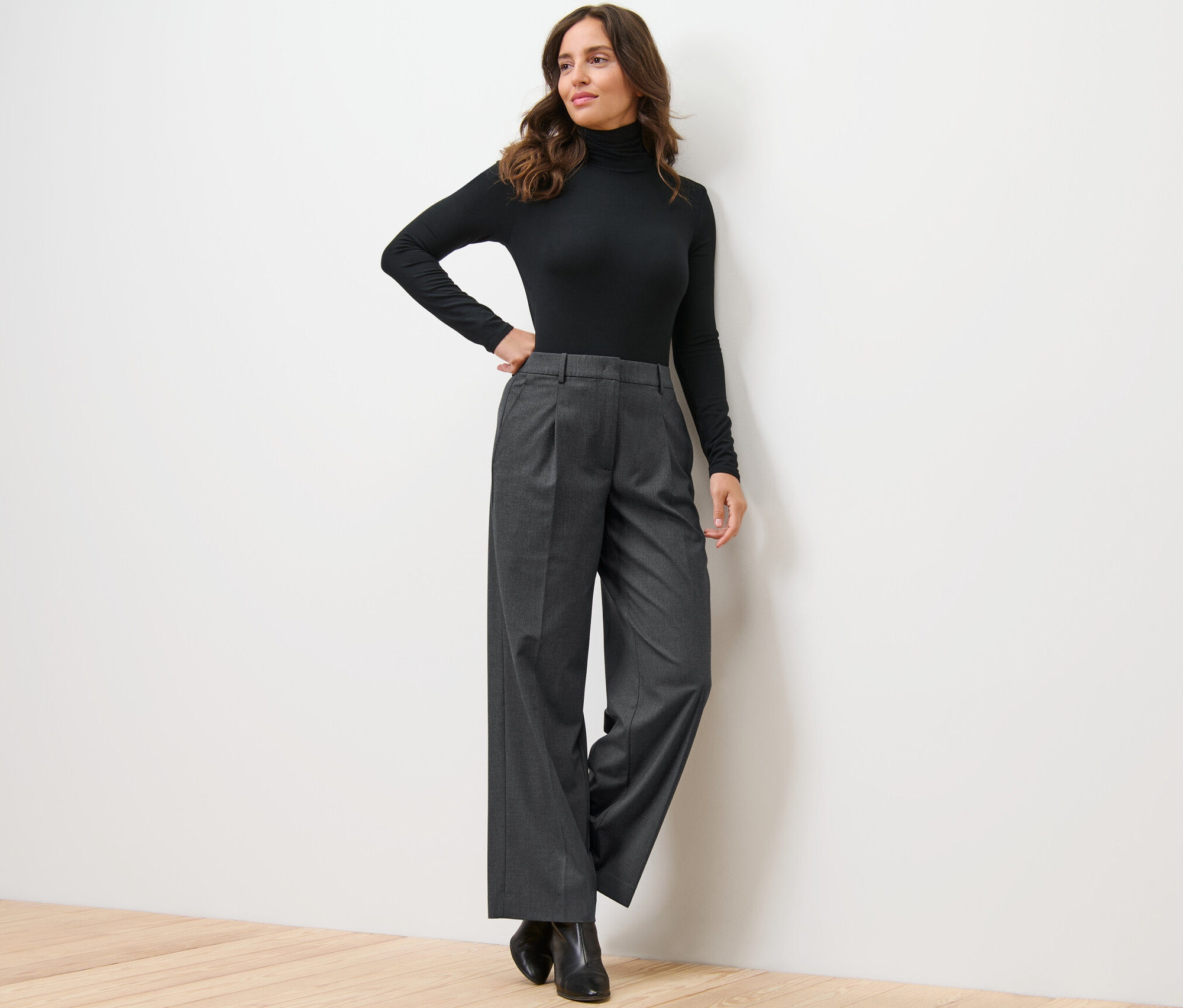 Une femme pose, appuyée contre un mur, portant un pull à col roulé noir et un pantalon tissé confortable.