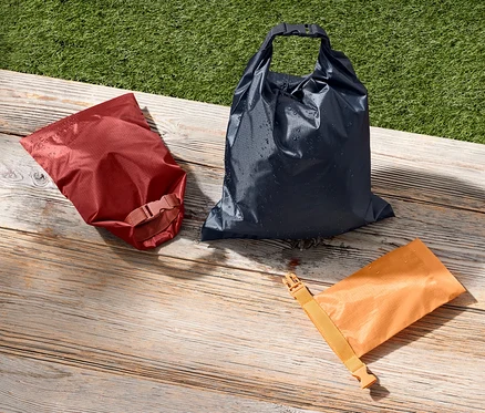 Trois sacs fourre-tout imperméables rouge, bleu et orange sont posés sur une surface en bois à côté d'une pelouse verte.