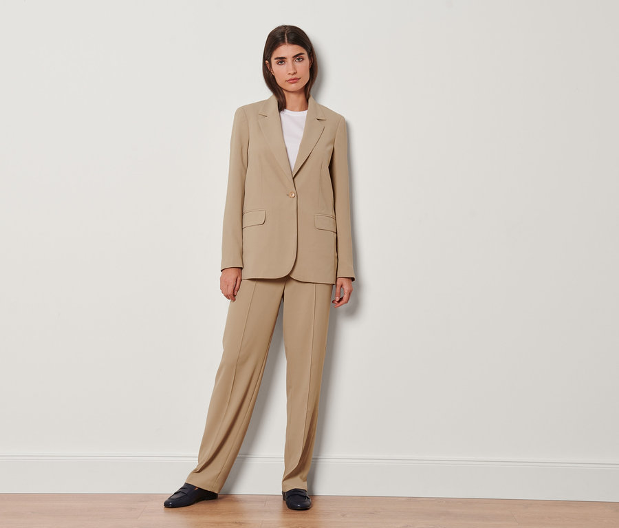 Une femme pose face à la caméra en blazer beige et pantalon tissé avec passepoil.