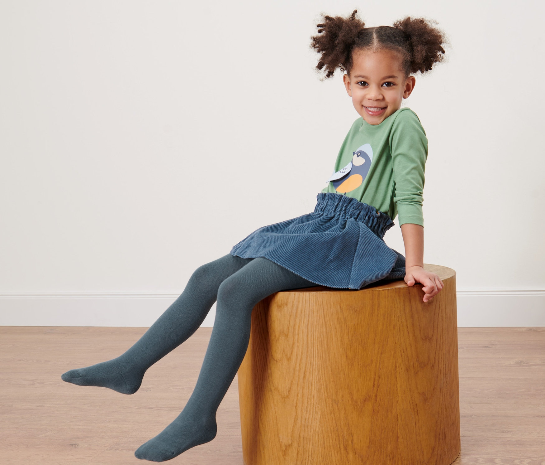 Une fillette souriante est assise sur un tabouret en bois et porte des collants bleus pour enfants.