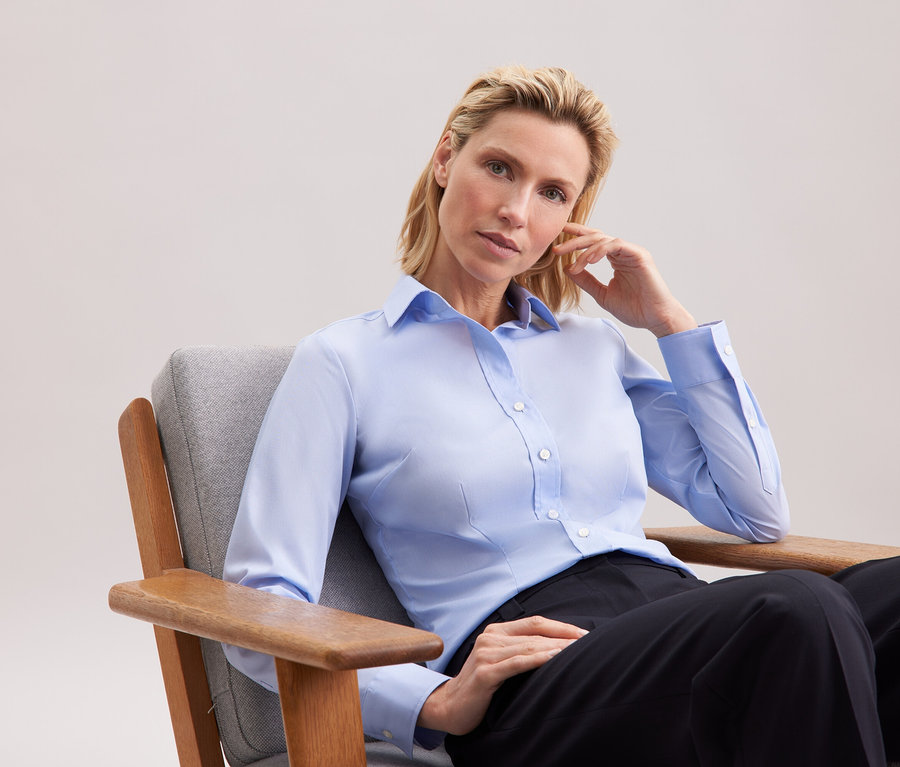 Une femme blonde est assise dans un fauteuil gris, portant une chemise bleu clair et un pantalon noir.