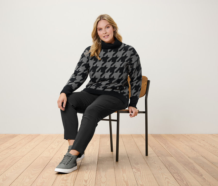 Une femme est assise sur une chaise, portant un pull pied-de-poule, un pantalon noir et des baskets grises.