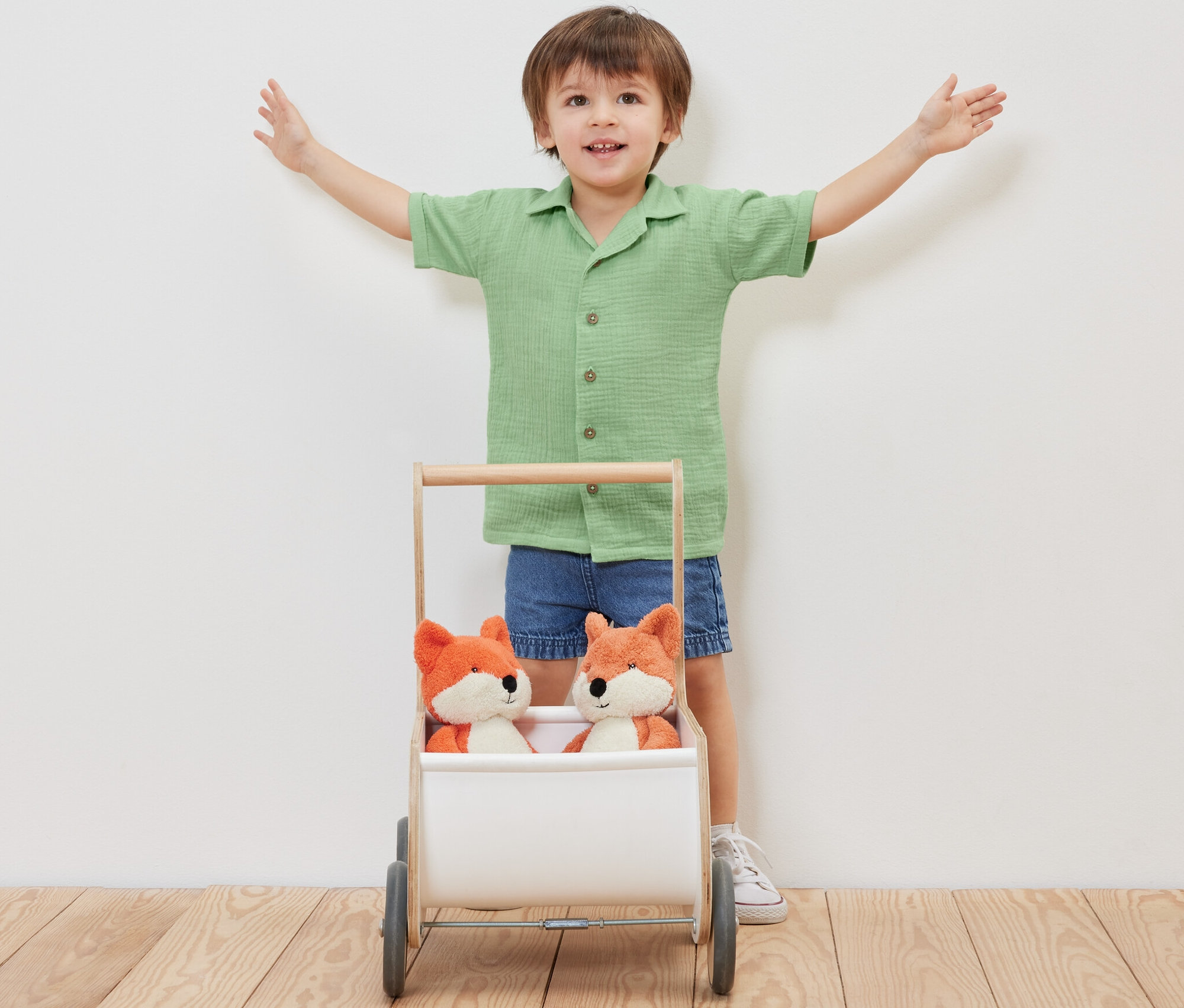 Un garçon se tient les bras écartés derrière un chariot contenant deux renards en peluche, portant une chemise en mousseline pour enfant avec col.