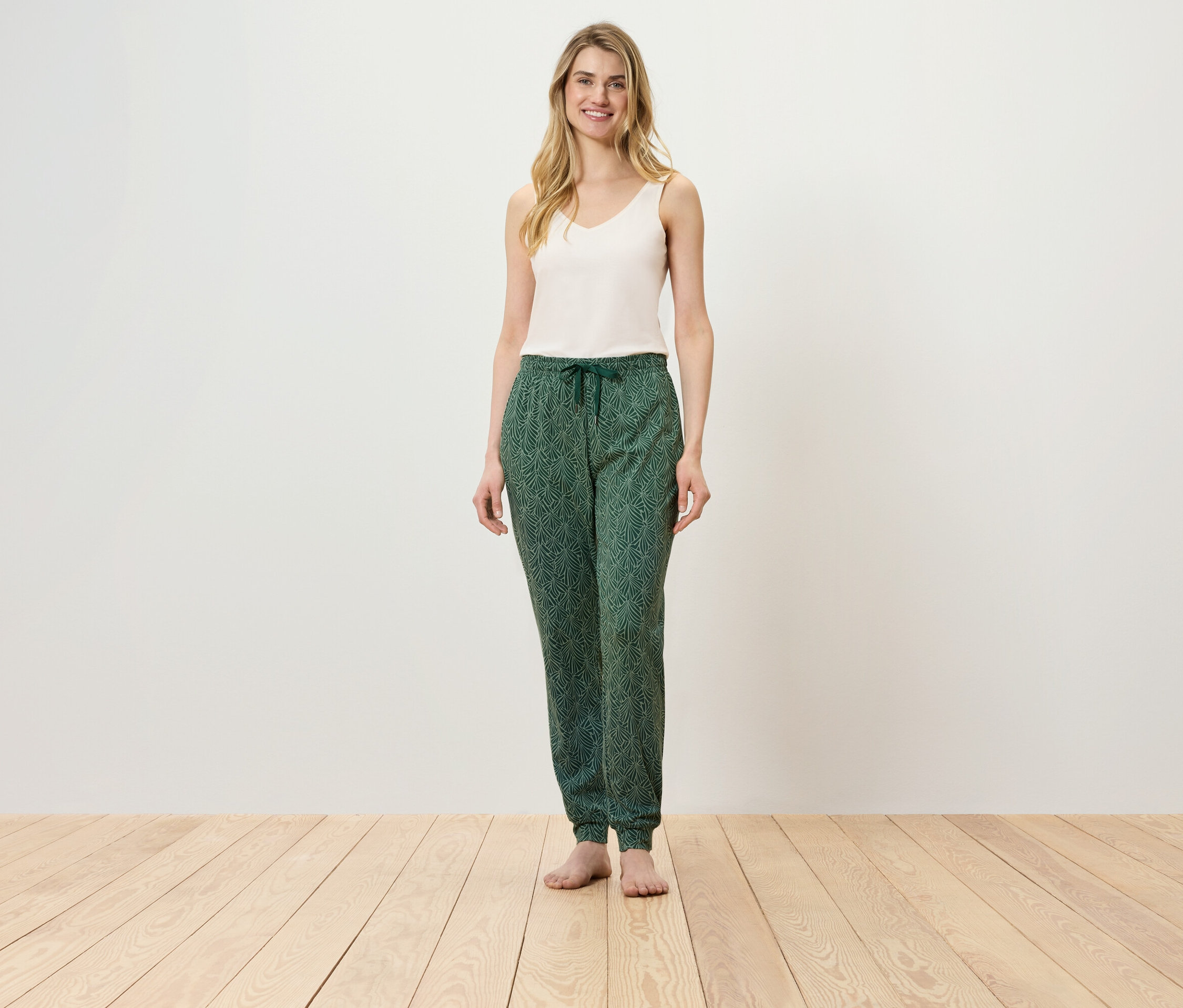 Une femme se tient sur un plancher en bois, portant un haut blanc et un pantalon vert.