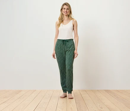 Une femme se tient sur un plancher en bois, portant un haut blanc et un pantalon vert.