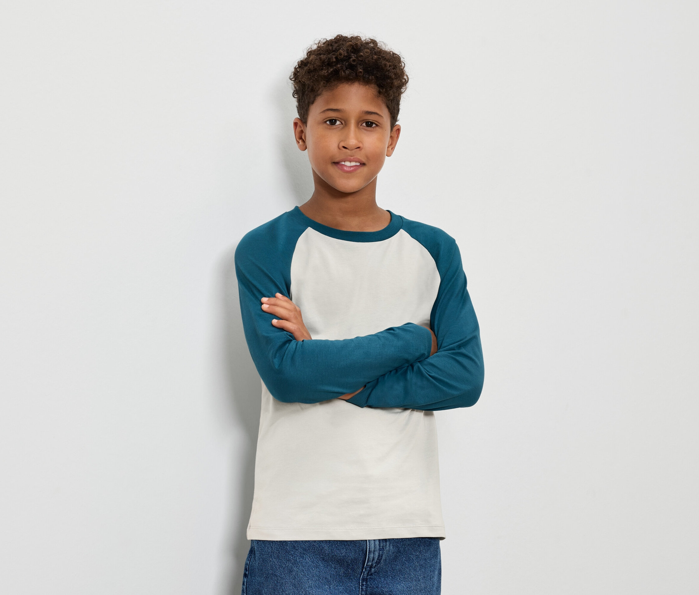 Un garçon aux cheveux bruns bouclés se tient debout, les bras croisés, portant un t-shirt à manches longues bleu-vert et un jean.