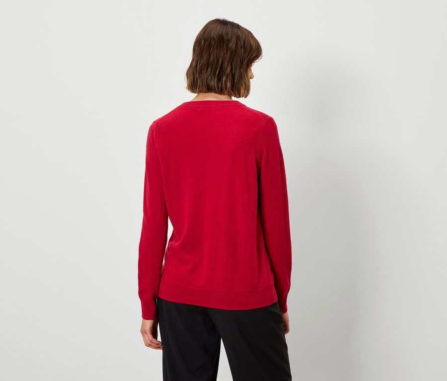 De dos, une femme aux cheveux bruns courts porte un pull rouge et un pantalon noir.