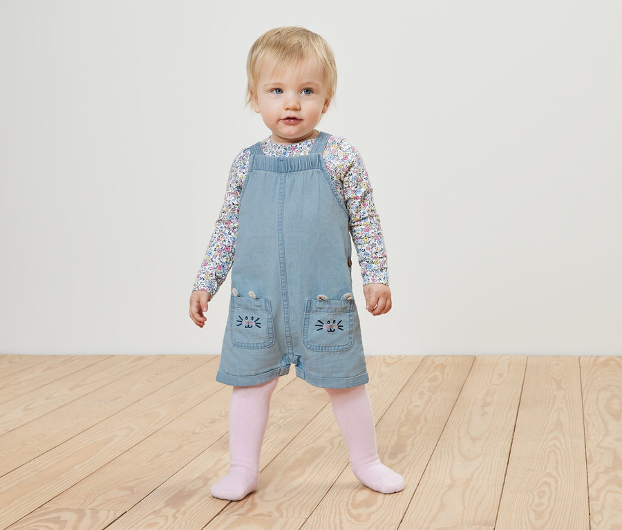 Un bébé portant une salopette en chambray pour enfant et un haut à fleurs se tient sur un plancher en bois.