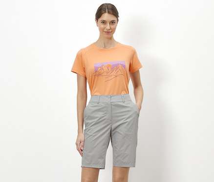 Une femme debout sur un fond blanc porte un t-shirt orange avec une illustration de montagnes et un short de cyclisme et de randonnée « Pro » gris.