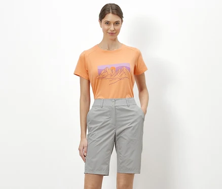 Une femme debout sur un fond blanc porte un t-shirt orange avec une illustration de montagnes et un short de cyclisme et de randonnée « Pro » gris.
