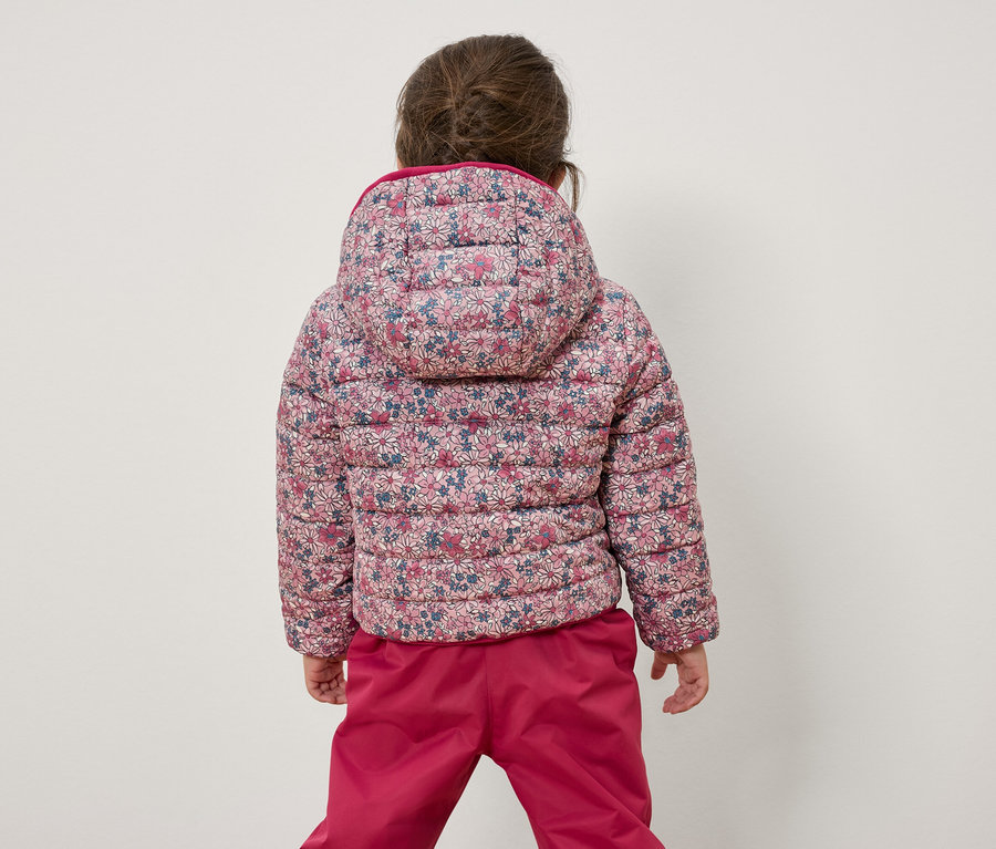 Une fillette, dos à la caméra, porte une veste à fleurs et un pantalon rose.