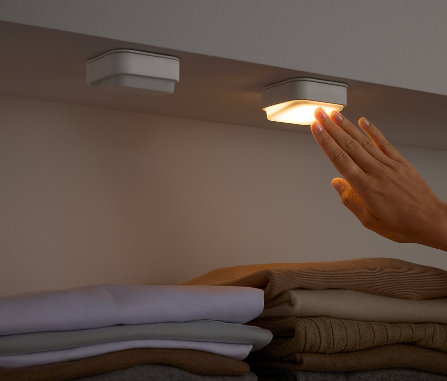 Une main touche une lumière carrée sous une étagère au-dessus de vêtements pliés.