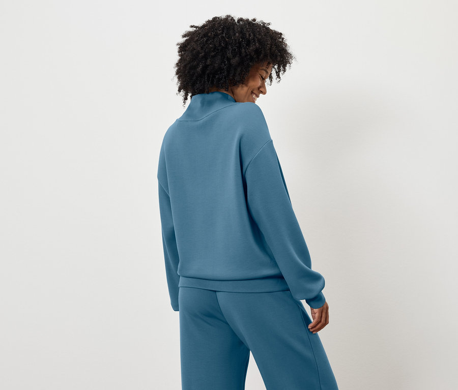 Vue de dos, une femme aux cheveux bouclés noirs porte un pull et un pantalon bleus.
