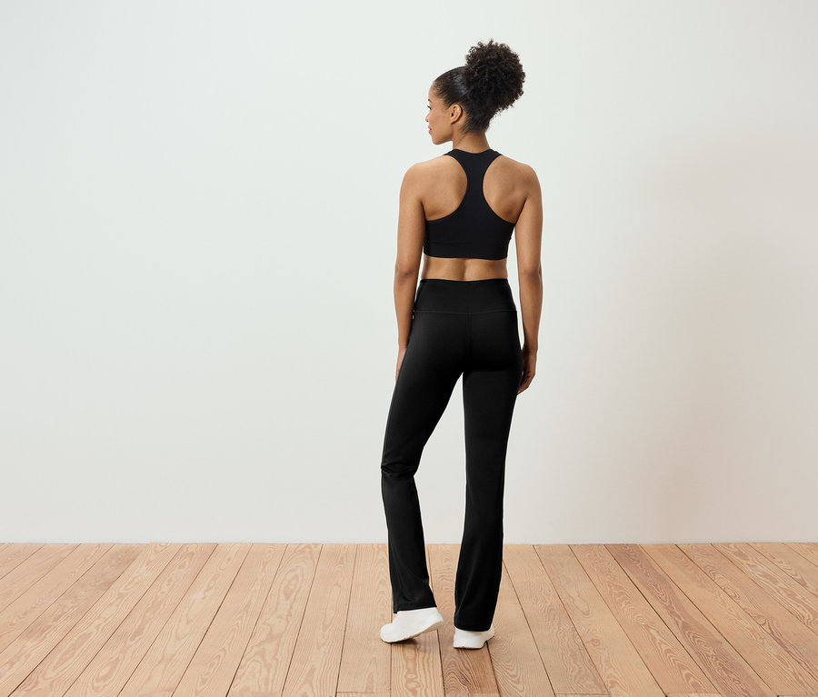 Vue de dos d'une femme aux cheveux relevés portant un soutien-gorge de sport noir et un pantalon noir. Elle se tient debout sur un plancher en bois.