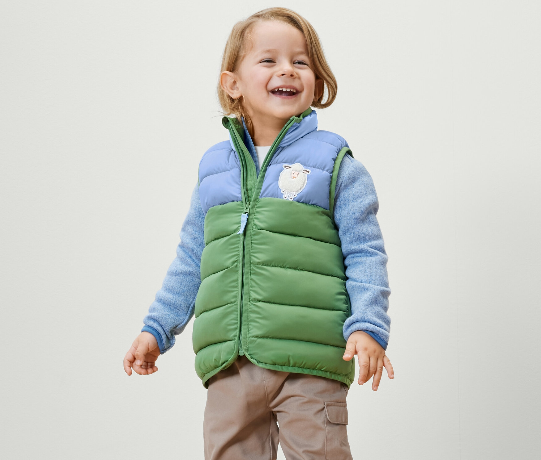 Un petit garçon souriant porte un gilet sans manches matelassé color block, un pull bleu et un pantalon.