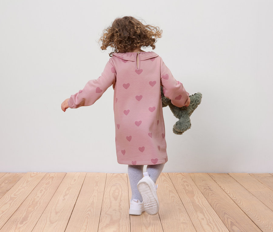 Une fillette aux cheveux bouclés, vêtue d'une robe rose à cœurs et de chaussures blanches, marche sur un plancher en bois et tient un ours en peluche vert.