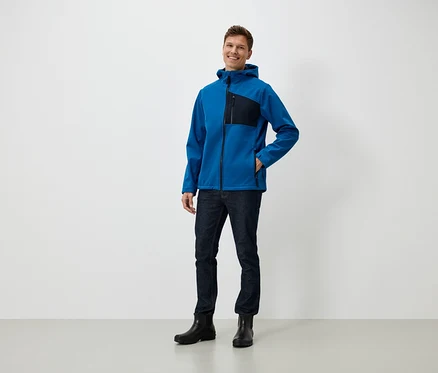 Un homme debout souriant porte une veste bleue à capuche, un jean et des bottes noires.
