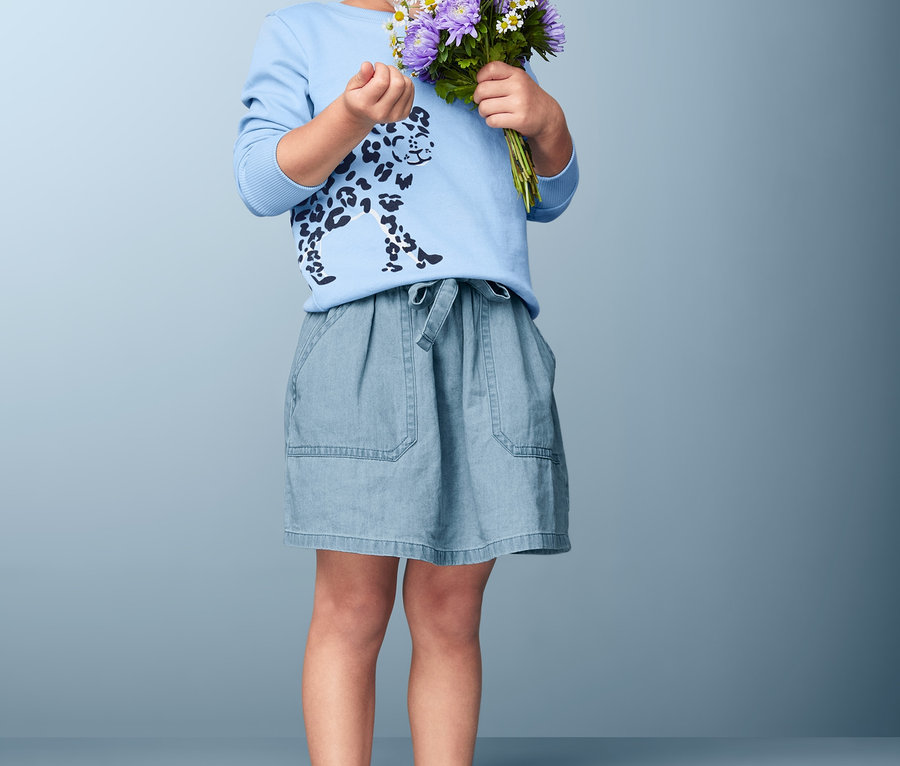 Une fillette bouclée sent un bouquet de fleurs violettes et blanches. Elle porte un t-shirt bleu à imprimé guépard et une jupe en denim de coton.
