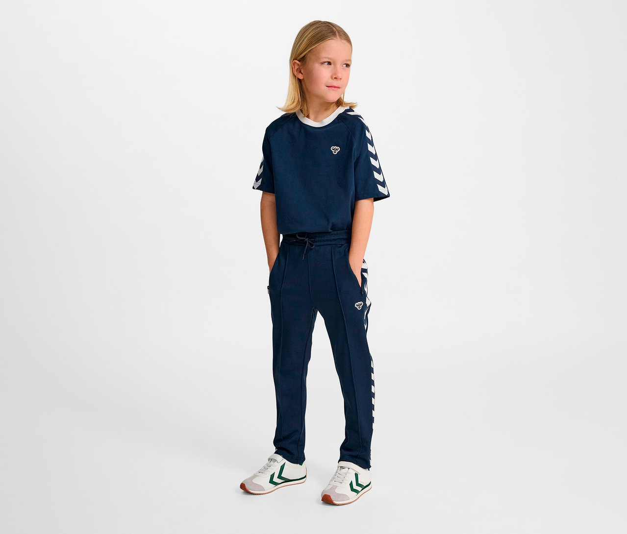 Une jeune fille pose dans un survêtement bleu marine HUMMEL HMLJR Archive Regular Poly Pants avec des détails blancs.